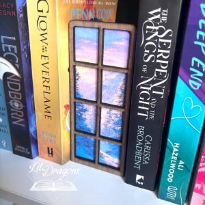 Puede incluir: Un marcapáginas de madera con diseño de ventana, que muestra una vista panorámica de un paisaje montañoso con árboles y un cielo colorido. El marcapáginas está colocado entre varios libros en una estantería.