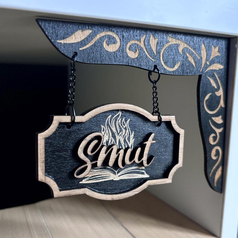 May include: A black wooden sign with a brown border hangs from a black chain. The sign has a white flame design and the word "Smut" written in a stylized font. The sign is hanging from a black wooden bracket with a decorative design.