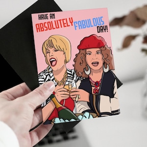 Puede incluir: Una tarjeta de felicitación rosa con una ilustración de dibujos animados de dos mujeres con cabello rubio y castaño, vestidas con ropa colorida, sosteniendo copas de champán. El texto de la tarjeta dice "Have an Absolutely Fabulous Day!"