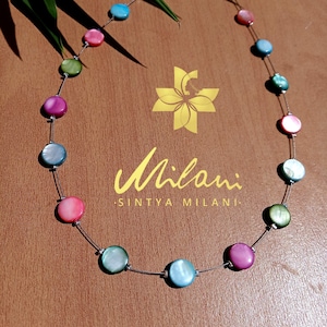 May include: A vibrant necklace with iridescent, circular beads in pink, blue, green, and purple, strung on a fine silver wire. The necklace is presented on a wooden surface with the text "Milani Sintya Milani".