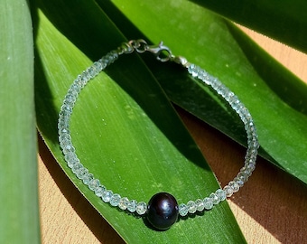 Op de afbeelding: Een delicate armband met kleine, lichtgroene kralen en een enkele, donkere, ronde kraal. De armband is geplaatst tegen een achtergrond van levendige groene bladeren en een bruin oppervlak, wat een natuurlijke en elegante uitstraling creëert.