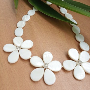 Puede incluir: Collar de conchas blancas con tres acentos de flores. Cada flor está hecha de cinco piezas de concha y tiene un strass transparente en el centro.