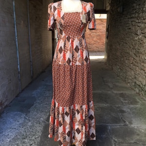Peut inclure: Une robe longue présentant un motif patchwork de formes géométriques et d'imprimés floraux dans des tons marron, orange et blanc. Le vêtement possède des manches courtes, une encolure carrée et une jupe à étages, présentée sur un mannequin.