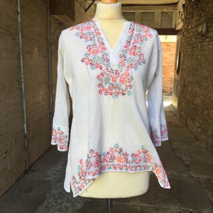 May include: A white long-sleeve tunic featuring a V-neckline with a button placket. The garment is decorated with colorful floral embroidery in shades of orange, teal, and purple around the neckline, the sleeve cuffs, and the bottom hem.