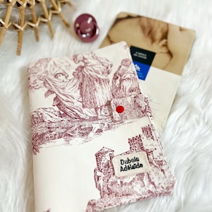 Puede incluir: Una cubierta de libro color crema con una ilustración burdeos de figuras y una escena de castillo. La cubierta tiene un cierre de botón rojo y una etiqueta que dice "Dubois Adélaïde". También se ven un chupete y un sobre vintage.