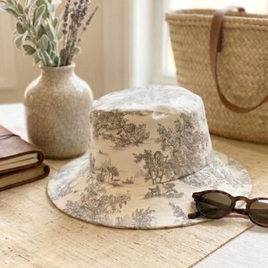 May include: A wide-brimmed hat with a cream and gray toile pattern, resting on a neutral-toned table runner. Tortoiseshell sunglasses are placed near the hat. A vase with greenery and a woven tote bag are in the background.