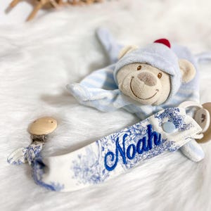 Peut inclure: Une couverture de sécurité en forme d'ours avec des rayures bleu clair et blanches, accompagnée d'une attache-sucette à motif floral bleu et blanc avec le prénom Noah brodé en bleu, dotée d'un clip en bois et d'un bouton-pression bleu clair.