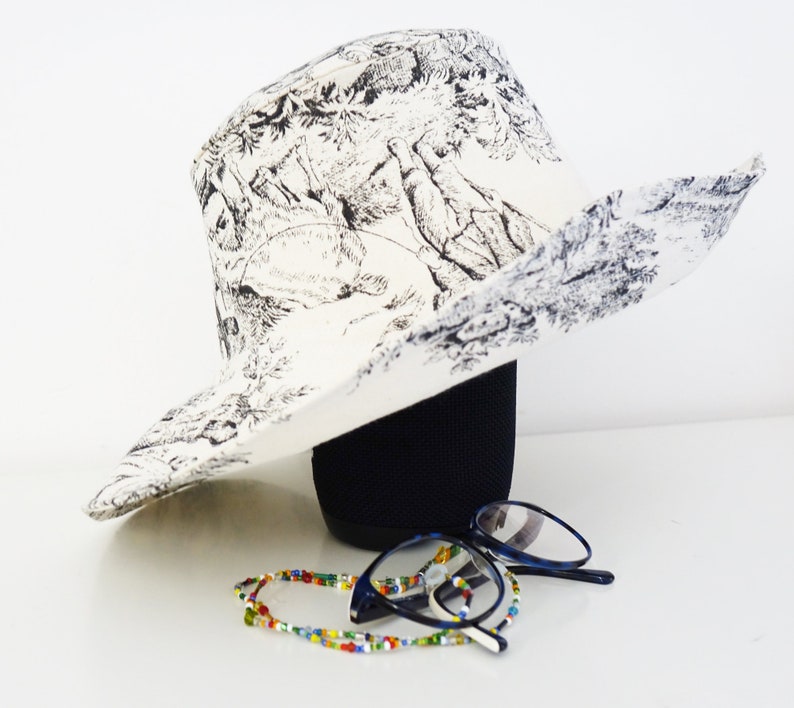 Grand chapeau à larges bords en Toile de Jouy blanche, fait main en France. Élégant et intemporel, il protège du soleil avec style. Doublure unie et surpiqûres raffinées sur la capeline pour une finition haut de gamme. Accessoire chic pour mariage, cérémonie, jardin ou plage. Idée cadeau femme, accessoire mode français, parfait pour la fête des mères, un anniversaire ou une escapade élégante sous le soleil