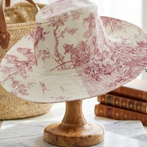 May include: A wide-brimmed hat with a cream-colored background and a burgundy toile pattern depicting a pastoral scene. The hat is displayed on a wooden hat stand. A straw bag and books are in the background.