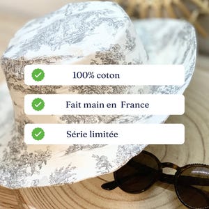 May include: A white and gray patterned sun hat with a wide brim. The hat is made of 100% cotton and is handmade in France. The hat is a limited edition. Tortoiseshell sunglasses rest on a wooden surface.