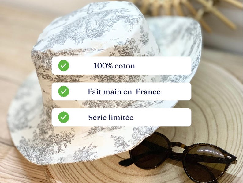 May include: A white and gray patterned sun hat with a wide brim. The hat is made of 100% cotton and is handmade in France. The hat is a limited edition. Tortoiseshell sunglasses rest on a wooden surface.