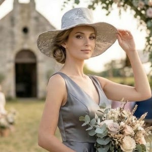 May include: A woman wearing a silver-gray wide-brimmed hat with a floral pattern and a matching V-neck dress. She holds a bouquet of flowers with roses and eucalyptus. The background features a blurred outdoor setting with a building.
