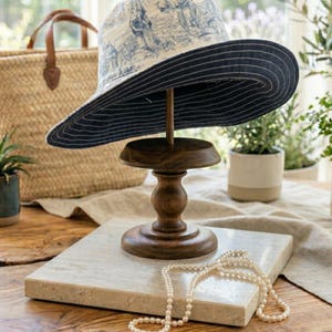May include: A wide-brimmed hat with a blue and white toile pattern, displayed on a wooden stand. The hat has a navy blue underside and is set on a decorative wooden pedestal. A pearl necklace rests on a marble tile.