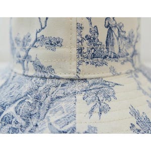 May include: Close-up of a bucket hat featuring a blue and white toile pattern. The fabric is a cream color with a repeating design of pastoral scenes. The hat has a structured brim and is made of a durable material.