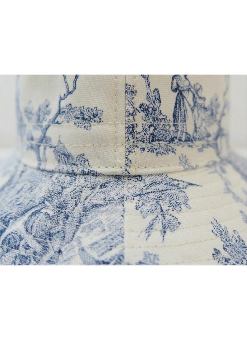 May include: Close-up of a bucket hat featuring a blue and white toile pattern. The fabric is a cream color with a repeating design of pastoral scenes. The hat has a structured brim and is made of a durable material.