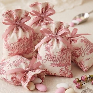 Peut inclure: Plusieurs petits sacs en tissu crème avec un motif de toile de Jouy rose et des rubans roses. Chaque sac est brodé du nom "Juliette". Certains sacs sont ouverts, révélant des bonbons roses et blancs.
