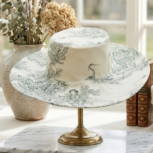 May include: A white sun hat with a wide brim, featuring a toile pattern in shades of green. The hat is displayed on a gold-colored stand. The toile design depicts a pastoral scene with figures and foliage.