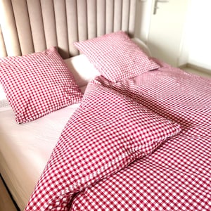 Puede incluir: Un juego de cama con un patrón de cuadros vichy en rojo y blanco en la funda nórdica y dos fundas de almohada cuadradas. La ropa de cama está dispuesta sobre una cama con un cabecero alto, tapizado en beige claro y con costuras verticales.