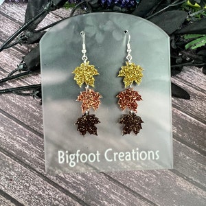 May include: A pair of dangle earrings featuring three layered glitter maple leaves in gold, copper, and brown colors. The earrings are displayed on a white card with the text "Bigfoot Creations".