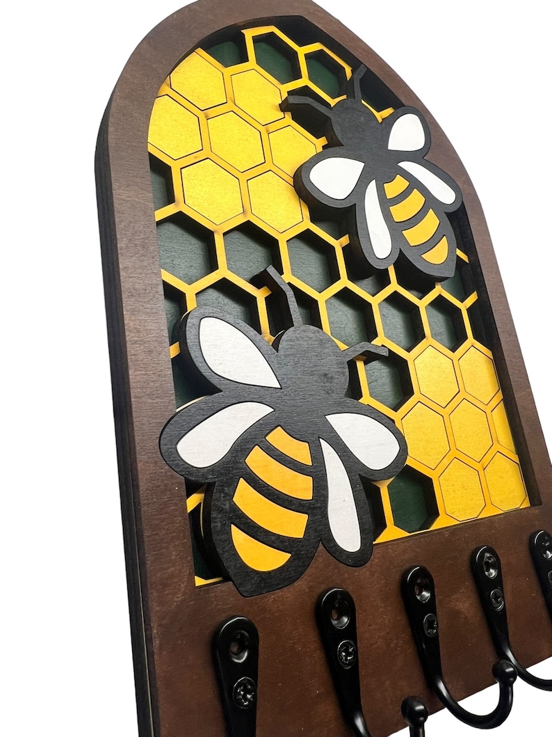 May include: Wooden key holder with a honeycomb design and two bees. The bees are black and yellow with white wings. There are five hooks on the bottom of the holder.