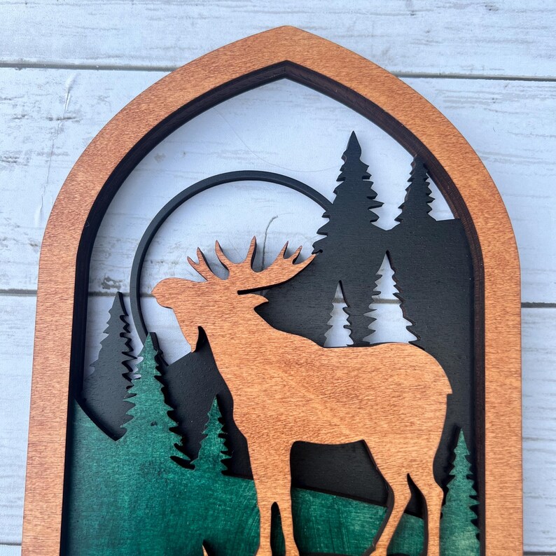 May include: A wooden wall hanging with a layered design featuring a moose silhouette in front of a full moon and a forest scene. The moose is brown, the moon is black, and the forest is green and black.