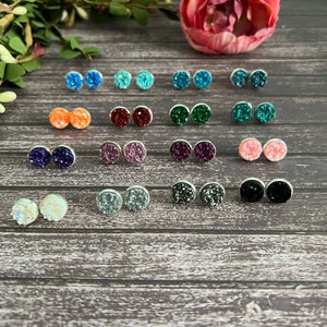 May include: Assortment of 12 pairs of small round stud earrings with various colors of glitter embedded in the resin. The earrings are displayed on a wooden surface.