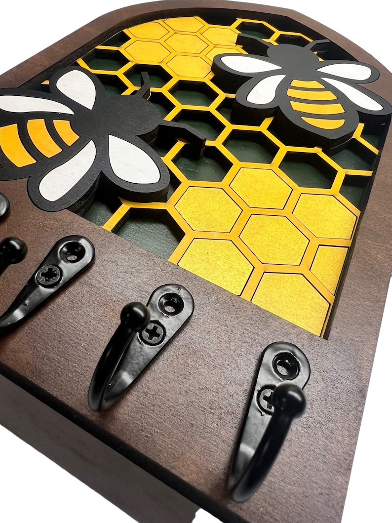 May include: Wooden key holder with three hooks, featuring a honeycomb design with two black and yellow bees. The key holder is brown with a black border.