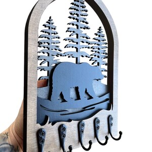 May include: Wooden key holder featuring a bear and pine tree design. The key holder has a grey frame with a black bear silhouette, pine trees, and five black metal hooks for keys. The key holder is held by a person.