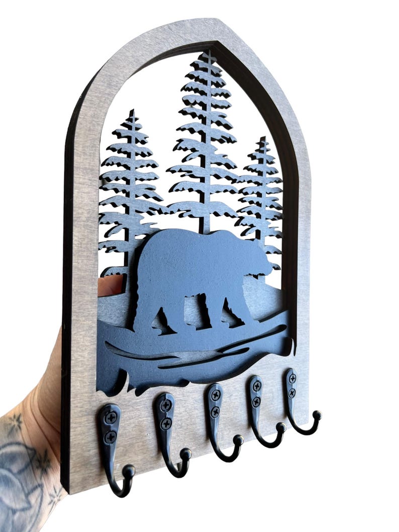 May include: Wooden key holder featuring a bear and pine tree design. The key holder has a grey frame with a black bear silhouette, pine trees, and five black metal hooks for keys. The key holder is held by a person.