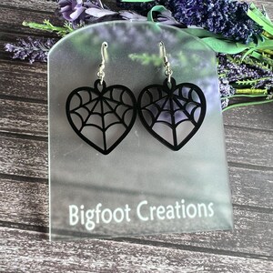 May include: Black heart-shaped earrings with a spiderweb design. The earrings are on a white display card with the text "Bigfoot Creations".