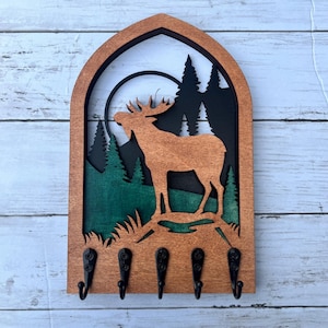 May include: Wooden key holder with a moose silhouette in a forest scene. The moose is brown, the trees are green, and the background is black. There are five hooks on the bottom of the key holder.