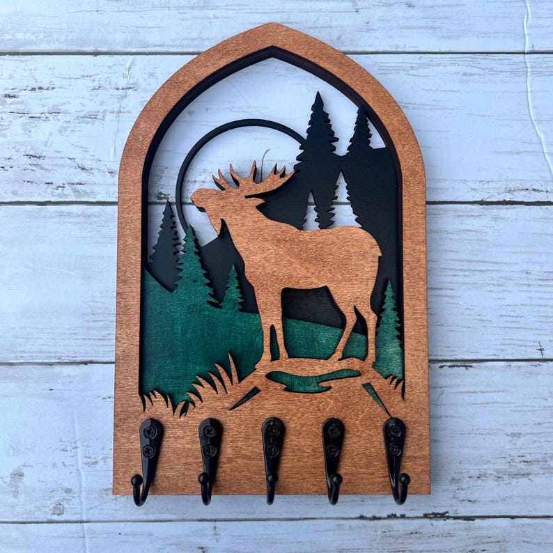 May include: Wooden key holder with a moose silhouette in a forest scene. The moose is brown, the trees are green, and the background is black. There are five hooks on the bottom of the key holder.