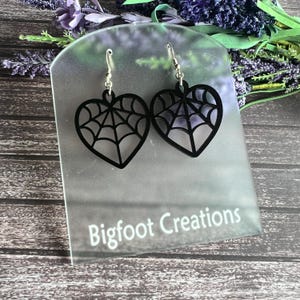 May include: Black heart-shaped earrings with a spiderweb design. The earrings are on a clear display stand with the text "Bigfoot Creations".