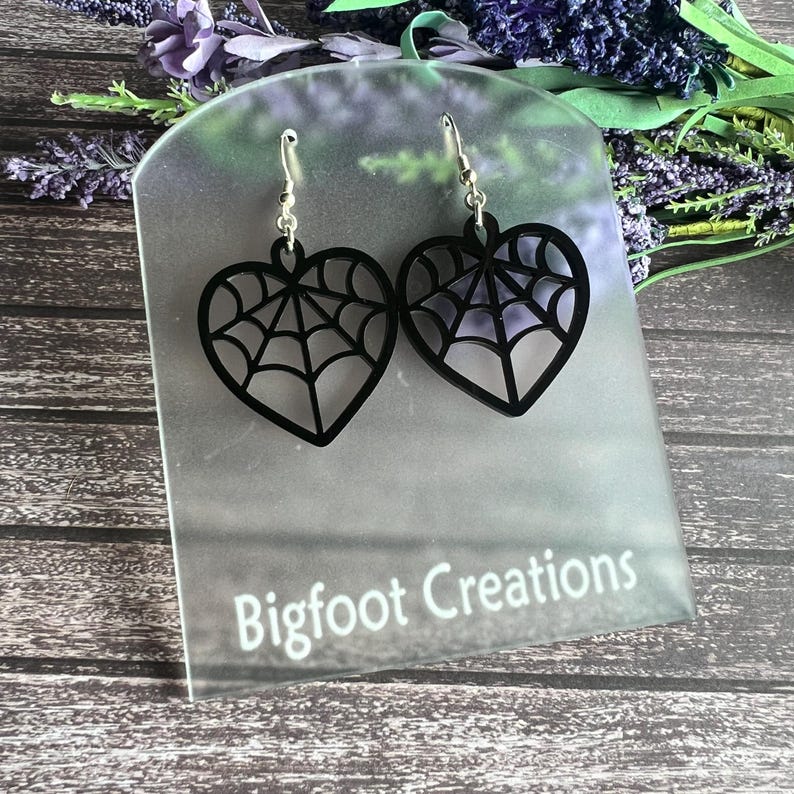 May include: Black heart-shaped earrings with a spiderweb design. The earrings are on a clear display stand with the text "Bigfoot Creations".