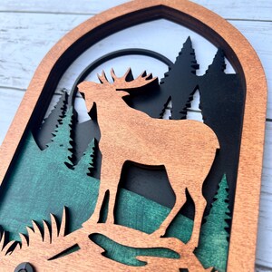 May include: A wooden wall hanging with a layered design featuring a moose silhouette in front of a forest scene. The moose is brown, the trees are black, and the background is green.