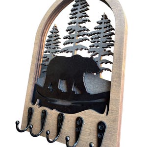 May include: Wooden key holder featuring a bear and pine tree silhouette design. The key holder has a dark brown wooden frame with a cutout of a bear walking in front of pine trees. It features five black metal hooks for keys or other small items.