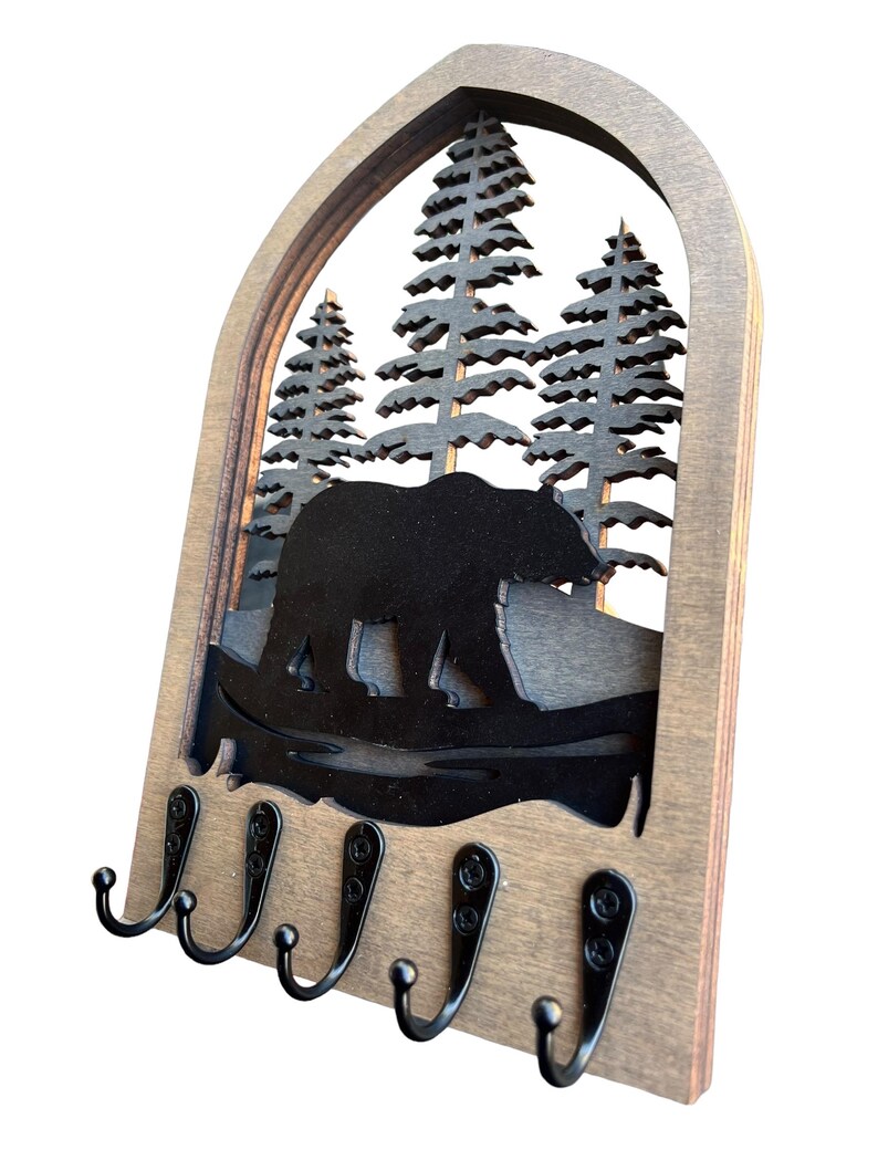 May include: Wooden key holder featuring a bear and pine tree silhouette design. The key holder has a dark brown wooden frame with a cutout of a bear walking in front of pine trees. It features five black metal hooks for keys or other small items.