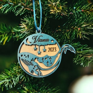 May include: A teal and wood-toned Christmas ornament with a whale design. The ornament features the name "Viana" and the year "2023". It is hanging from a blue string and set against a backdrop of green pine needles.