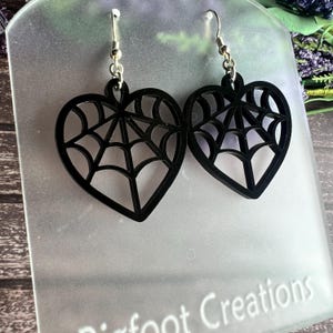 May include: Black heart-shaped earrings with a spiderweb design. The earrings are made of acrylic and have silver hooks.