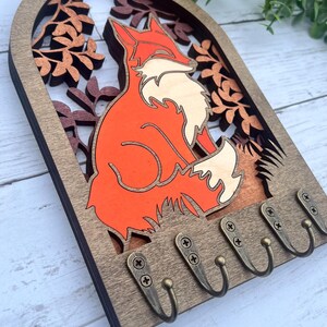 May include: Wooden key holder with a fox silhouette in orange and white, surrounded by brown leaves. The key holder has five hooks for hanging keys.