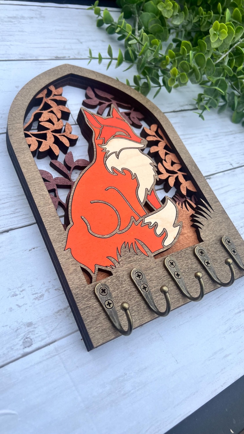 May include: Wooden key holder with a fox silhouette in orange and white, surrounded by brown leaves. The key holder has five hooks for hanging keys.