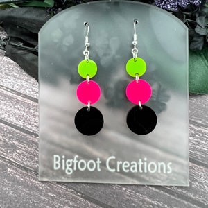 May include: A pair of dangle earrings with three round discs. The top disc is neon green, the middle disc is hot pink, and the bottom disc is black.
