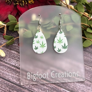 May include: A pair of teardrop-shaped earrings with a white background and a green marijuana leaf pattern. The earrings are hanging from silver hooks and are displayed on a white card with the text "Bigfoot Creations".