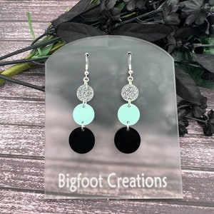May include: A pair of dangle earrings with three round discs. The top disc is silver with glitter, the middle disc is light blue, and the bottom disc is black.