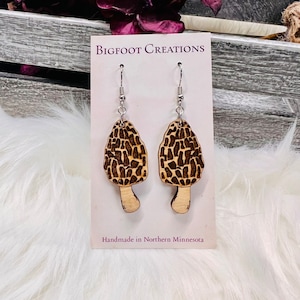 May include: A pair of wooden mushroom earrings with a brown and black pattern. The earrings are on a white card with the text "BIGFOOT CREATIONS" and "Handmade in Northern Minnesota".