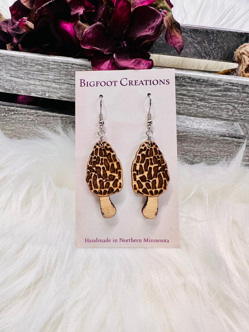 May include: A pair of wooden mushroom earrings with a brown and black pattern. The earrings are on a white card with the text "BIGFOOT CREATIONS" and "Handmade in Northern Minnesota".