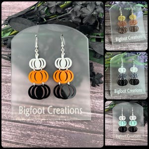 May include: Three-tiered pumpkin dangle earrings with white, orange, and black pumpkin shapes. The earrings are displayed on a white card with the text "Bigfoot Creations".