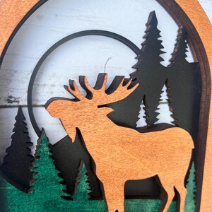 May include: A wooden cutout of a moose standing in a forest with a full moon behind it. The moose is brown and the trees are green and black.