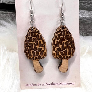 May include: A pair of wooden earrings shaped like morel mushrooms. The earrings are brown with a dark brown pattern and have silver hooks. The earrings are handmade in Northern Minnesota. "BIGFOOT CREATIONS" is printed on the card.