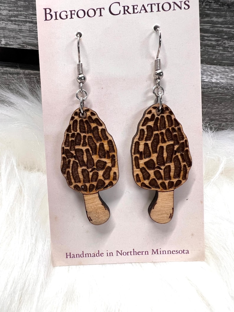 May include: A pair of wooden earrings shaped like morel mushrooms. The earrings are brown with a dark brown pattern and have silver hooks. The earrings are handmade in Northern Minnesota. "BIGFOOT CREATIONS" is printed on the card.